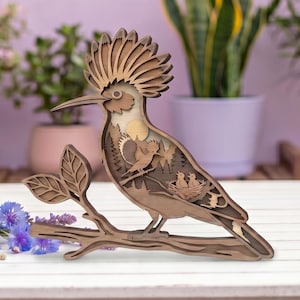 Può includere: Una scultura in legno multistrato raffigurante un'upupa appollaiata su un ramo. L'uccello presenta una cresta prominente e un design intagliato che rivela una scena boschiva con un uccellino, un sole e due pulcini nel nido all'interno del corpo.