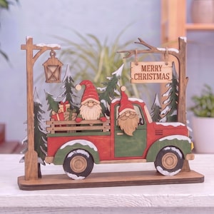 Pode incluir: Uma decoração de mesa em madeira com um camião vermelho e verde a transportar presentes e dois gnomos. A cena inclui pinheiros cobertos de neve, um candeeiro de rua e uma placa suspensa com o texto MERRY CHRISTMAS em letras pretas.