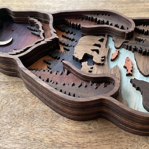 K&ouml;nnte beinhalten: Ein mehrschichtiges Kunstwerk in B&auml;renform aus Holz. Das Kunstwerk zeigt eine Waldszene mit einem B&auml;ren, Fischen, B&auml;umen und einem Mond. Es besteht aus verschiedenen Braunt&ouml;nen mit blauen und orangen Akzenten.