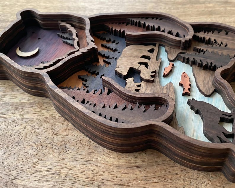 K&ouml;nnte beinhalten: Ein mehrschichtiges Kunstwerk in B&auml;renform aus Holz. Das Kunstwerk zeigt eine Waldszene mit einem B&auml;ren, Fischen, B&auml;umen und einem Mond. Es besteht aus verschiedenen Braunt&ouml;nen mit blauen und orangen Akzenten.