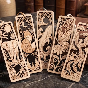 Op de afbeelding: Vijf rechthoekige houten bladwijzers met gedetailleerde lasergesneden ontwerpen van een olifant, een vos, een walvis, een vlinder met bloem en een feniks. Elke bladwijzer heeft een donkerbruine achtergrond, lichte houten uitsparingen en een kwastje.