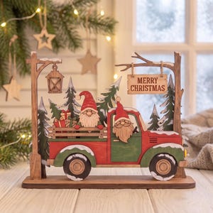 May include: A wooden tabletop decoration featuring a red and green truck carrying gnomes and gifts. The scene includes a street lamp, evergreen trees, and a hanging sign that reads MERRY CHRISTMAS. The design is layered with snow-dusted details.