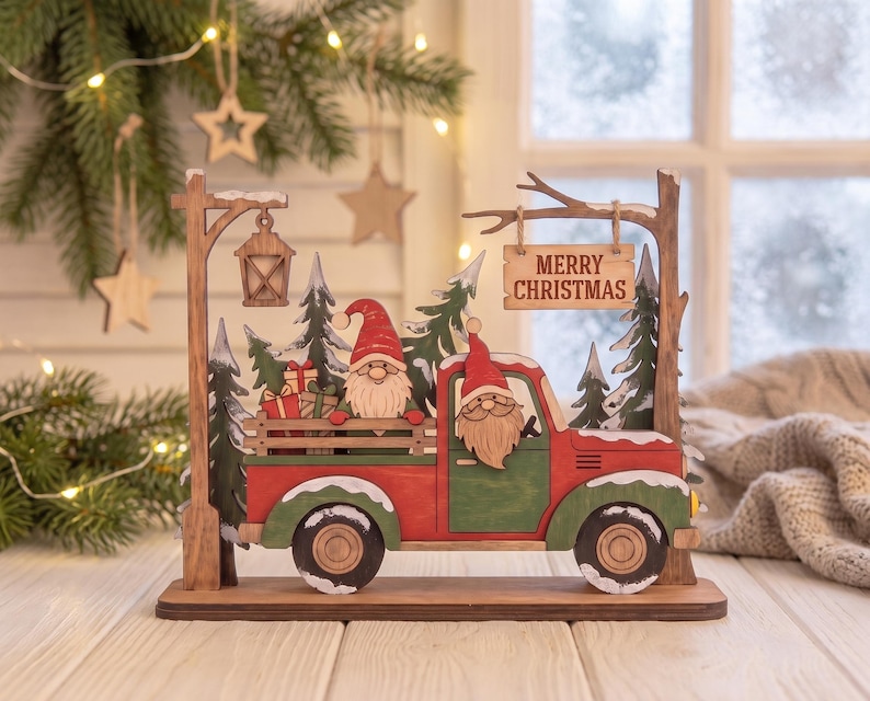 May include: A wooden tabletop decoration featuring a red and green truck carrying gnomes and gifts. The scene includes a street lamp, evergreen trees, and a hanging sign that reads MERRY CHRISTMAS. The design is layered with snow-dusted details.