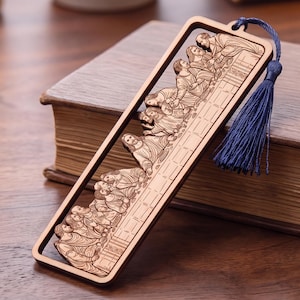 Peut inclure: Un marque-page rectangulaire en bois présentant une découpe laser représentant La Cène. Le motif complexe montre des personnages assis à une longue table. Un pompon en tissu bleu foncé est attaché au coin supérieur droit par un petit trou.