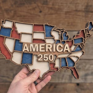 Może przedstawiać: Drewniana mapa Stanów Zjednoczonych z czerwonymi, białymi i niebieskimi sekcjami. Słowo "AMERICA" jest wyświetlane wypukłymi literami, z "250" poniżej. Mapa jest trzymana na tle drewnianym.