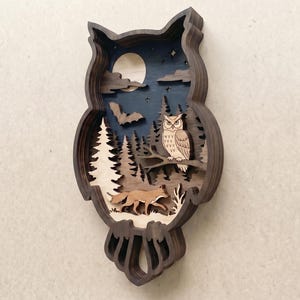 Op de afbeelding: Een meerlaags houten wanddecoratie in de vorm van een uil-silhouet. Het interieur toont een nachtsc&egrave;ne met een lichte maan, een vliegende vleermuis, een dennenbos, een zittende uil en een vos die op de grond beneden loopt.