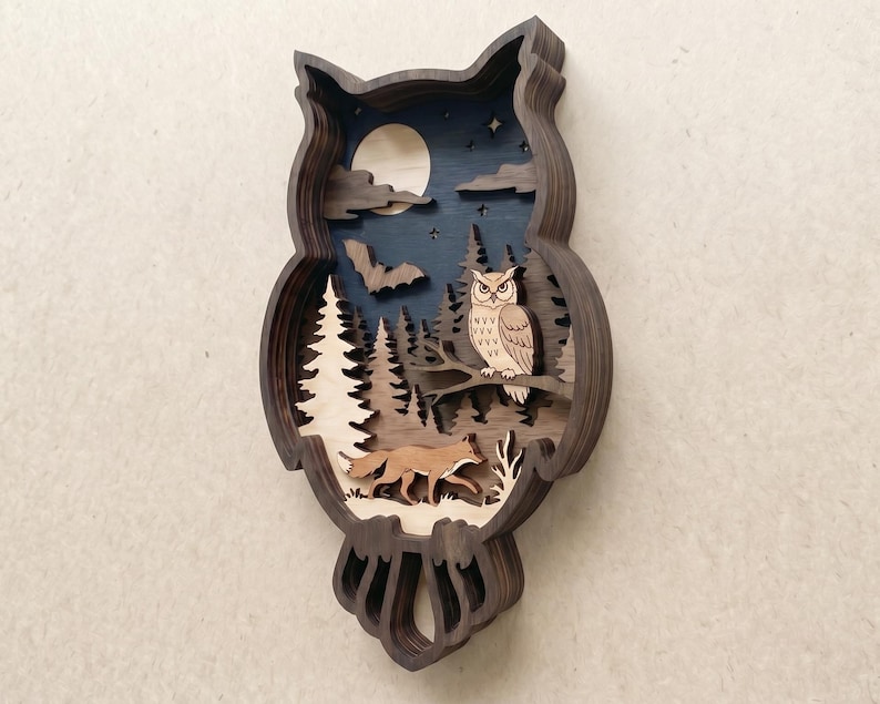 Op de afbeelding: Een meerlaags houten wanddecoratie in de vorm van een uil-silhouet. Het interieur toont een nachtsc&egrave;ne met een lichte maan, een vliegende vleermuis, een dennenbos, een zittende uil en een vos die op de grond beneden loopt.