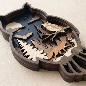 Op de afbeelding: Een meerlaags houten wanddecoratie in de vorm van een uil, met een donkerblauwe nachtelijke hemel met een maan, sterren en een vleermuis. De sc&egrave;ne bevat een lichte uil op een tak boven een bos met een vos die tussen dennenbomen loopt.