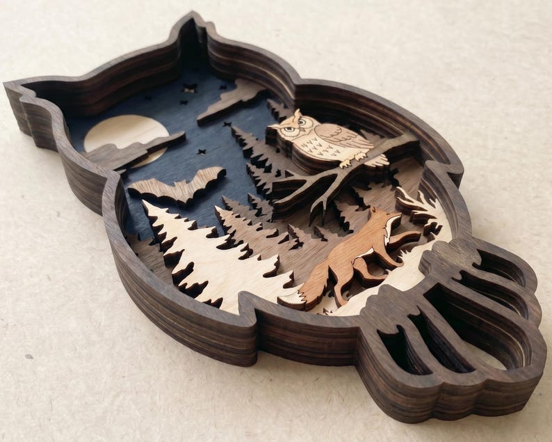 Op de afbeelding: Een meerlaags houten wanddecoratie in de vorm van een uil, met een donkerblauwe nachtelijke hemel met een maan, sterren en een vleermuis. De sc&egrave;ne bevat een lichte uil op een tak boven een bos met een vos die tussen dennenbomen loopt.