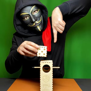 以下が含まれることがあります： サイコロのデザインの木製手品箱と、ドットマークの付いた別の木製サイコロ。 「LASER CUT FILES」と「Tricks for Kids and adults」の文字が見えます。 画像の人物は黒いマスクとフード付きスウェットを着ています。