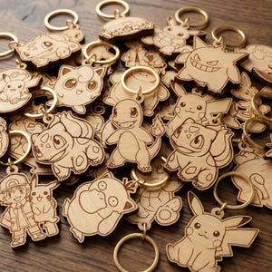 Peut inclure: Une collection de porte-clés en bois clair présentant des illustrations gravées au laser de diverses créatures et personnages animés, dont un Salamèche central, chacun attaché à un anneau brisé en métal doré.