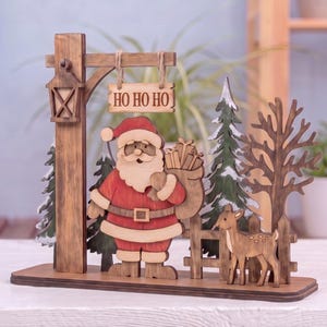 Puede incluir: Una decoración navideña de mesa hecha de madera que presenta una figura recortada de Santa Claus con un saco de regalos, un ciervo pequeño, árboles de hoja perenne, un árbol sin hojas, una valla y un letrero colgante que dice HO HO HO.