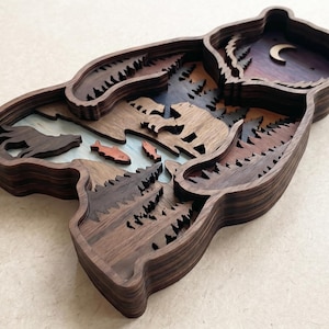 K&ouml;nnte beinhalten: Ein mehrschichtiges Holz-Kunstwerk in B&auml;renform. Das Kunstwerk zeigt eine Waldszene mit Fluss, Fischen, B&auml;umen und einer B&auml;rensilhouette. Die oberste Schicht zeigt einen Nachthimmel mit einem Halbmond. Das Holz hat eine dunkelbraune Oberfl&auml;che.