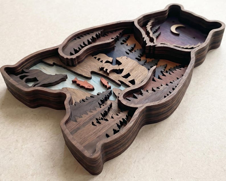 K&ouml;nnte beinhalten: Ein mehrschichtiges Holz-Kunstwerk in B&auml;renform. Das Kunstwerk zeigt eine Waldszene mit Fluss, Fischen, B&auml;umen und einer B&auml;rensilhouette. Die oberste Schicht zeigt einen Nachthimmel mit einem Halbmond. Das Holz hat eine dunkelbraune Oberfl&auml;che.