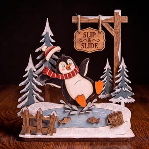 May include: A wooden decorative display featuring a penguin wearing a red and white striped scarf and hat, sliding on a blue ice patch. The scene includes evergreen trees, a wooden fence, a bucket, two fish, and a sign that reads SLIP & SLIDE.