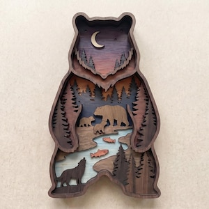Puede incluir: Una pieza de arte mural en forma de oso de madera en capas. El diseño presenta una escena de bosque con una familia de osos, un lobo, peces y árboles. La paleta de colores incluye tonos de marrón, azul, naranja y morado, con una luna creciente en la parte superior.