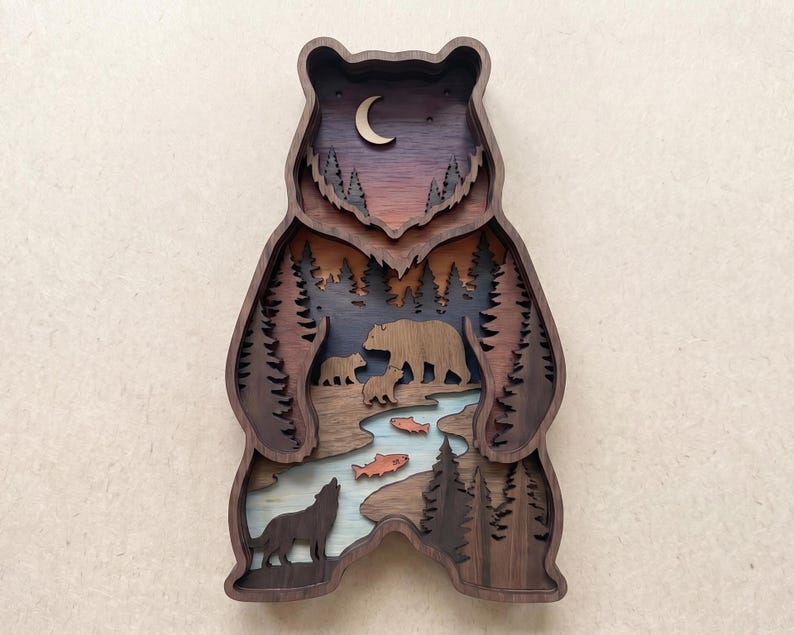 K&ouml;nnte beinhalten: Ein mehrschichtiges Wandkunstwerk in B&auml;renform aus Holz. Das Design zeigt eine Waldszene mit einer B&auml;renfamilie, einem Wolf, Fischen und B&auml;umen. Die Farbpalette umfasst Braunt&ouml;ne, Blau, Orange und Lila, mit einem Halbmond oben.