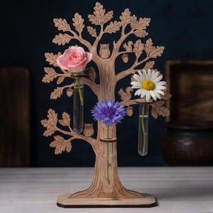 May include: A laser-cut wooden tree display featuring three clear glass test tube vases. The tree structure includes engraved details of owls, squirrels, and acorns. A pink rose, a blue cornflower, and a white daisy are placed in the tubes.