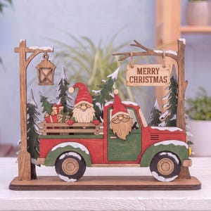 May include: A wooden decorative display featuring a red and green truck with two gnomes in the cab and bed, carrying wrapped gifts. The scene includes evergreen trees, a hanging lantern, and a sign that reads MERRY CHRISTMAS in black letters.