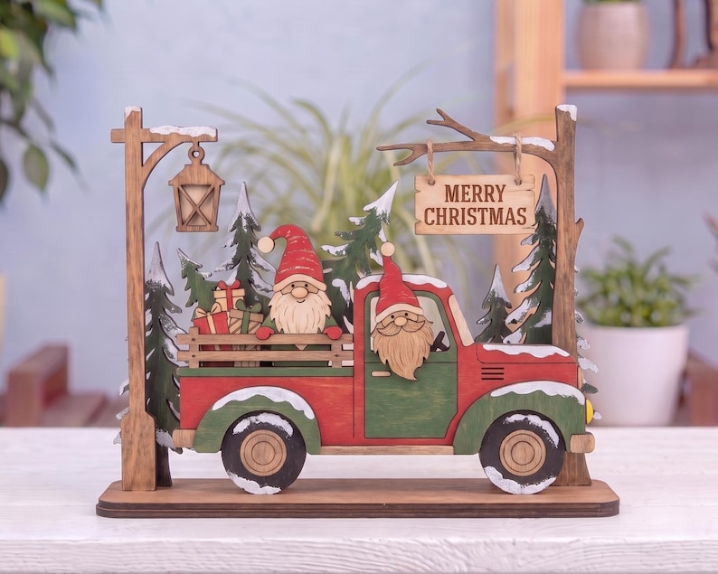 May include: A wooden decorative display featuring a red and green truck with two gnomes in the cab and bed, carrying wrapped gifts. The scene includes evergreen trees, a hanging lantern, and a sign that reads MERRY CHRISTMAS in black letters.