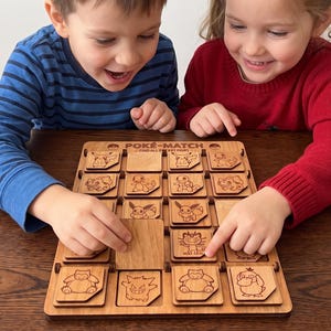 May include: A wooden memory matching game board featuring the text POKE-MATCH FIND ALL POCKET PAIRS. The board contains a grid of square wooden tiles, each engraved with various cartoon creature illustrations, with some tiles flipped over to reveal blank surfaces.