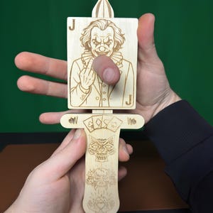 Peut inclure: Un jouet de tour de magie en bois découpé au laser avec un motif de carte Joker. Le jouet a une poignée avec des symboles de cartes à jouer, une tête de mort et le texte "Tricks for kids and adults" et "Laser Cut Files".