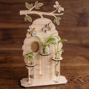 May include: A wooden bee-themed propagation station with a honeycomb design. It features three glass tubes for plants, with decorative wooden bees and leaves. The station is made of light-colored wood and is designed to hold small plants.