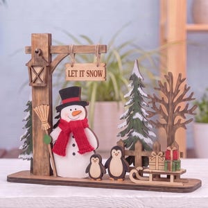 Könnte beinhalten: Eine winterliche Holzdekoration mit einem Schneemann mit rotem Schal und schwarzem Hut, zwei kleinen Pinguinen, einer Laterne, einem Schild mit der Aufschrift LET IT SNOW, Tannenbäumen, einem kahlen Baum, einem Holzzaun und einem Schlitten.