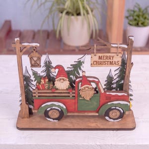 May include: A wooden tabletop decoration featuring a red and green truck with two gnomes, one in the driver seat and one in the truck bed with gifts. The scene includes green pine trees, a hanging lantern, and a sign that reads MERRY CHRISTMAS.