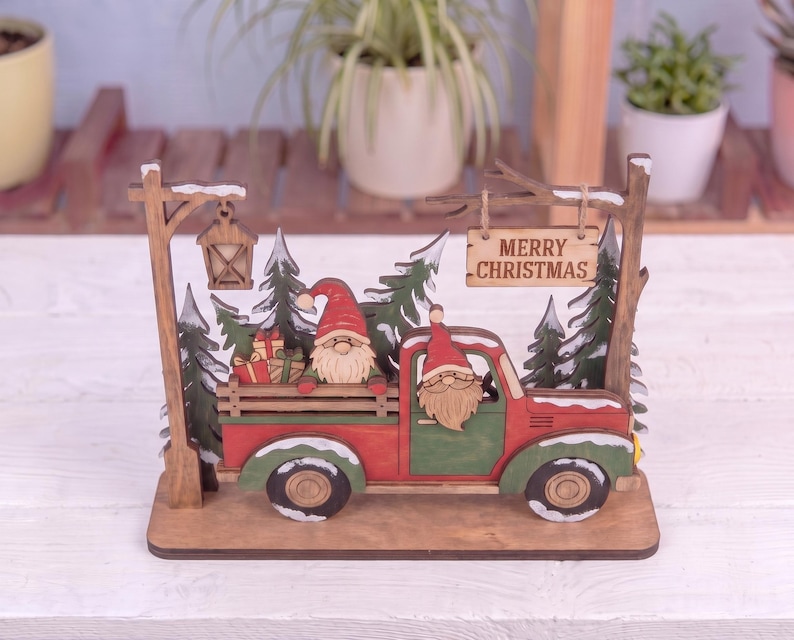 May include: A wooden tabletop decoration featuring a red and green truck with two gnomes, one in the driver seat and one in the truck bed with gifts. The scene includes green pine trees, a hanging lantern, and a sign that reads MERRY CHRISTMAS.