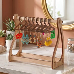 May include: A light-colored wooden earring display stand featuring a horizontal bar with several small, dark brown wooden hangers. Various pairs of earrings, including floral, crescent moon, tassel, and geometric designs, hang from the hangers.