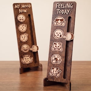 May include: Two vertical dark wood mood trackers with stands. The left board features the text "MY MOOD NOW:" and five emoji icons. The right board features the text "FEELING TODAY:" and five emoji icons. Each board has a vertical slot with a sliding pointer.