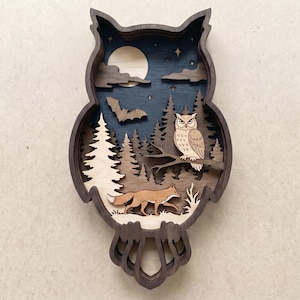 May include: A layered wooden owl-shaped wall art piece. The design features a forest scene with an owl perched on a branch, a fox, trees, a moon, and a bat. The color palette includes shades of brown, blue, and white.