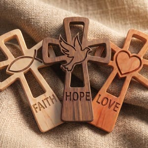 May include: Three wooden crosses with cut-out designs. The left cross is light wood with an ichthys symbol and the word FAITH. The center cross is dark wood with a dove symbol and the word HOPE. The right cross is medium wood with a heart and the word LOVE.