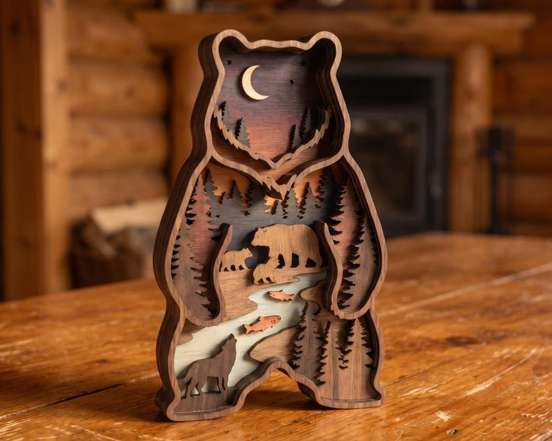 K&ouml;nnte beinhalten: Mehrschichtige Holzskulptur eines B&auml;ren mit einer Waldszene. Die Skulptur zeigt eine B&auml;renfamilie, einen Wolf, Fische und B&auml;ume. Der Hintergrund zeigt einen Sonnenuntergang und einen Mond. Hergestellt aus verschiedenen Braunt&ouml;nen.