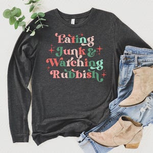 May include: A charcoal grey long-sleeved top featuring the phrase "Eating Junk & Watching Rubbish" in a retro font. The shirt is styled with blue denim jeans and beige ankle boots, a relaxed ensemble.