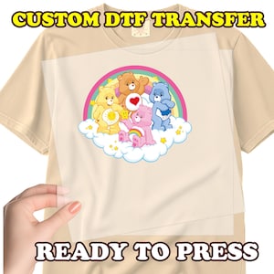 May include: A beige t-shirt with a DTF transfer featuring Care Bears on a cloud with a rainbow. The transfer is ready to press. The text "CUSTOM DTF TRANSFER" and "READY TO PRESS" are displayed in the image.