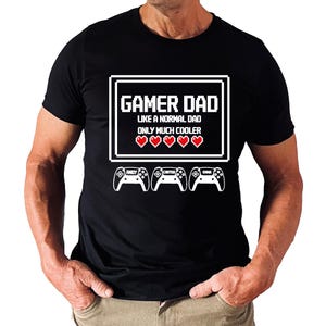 May include: Black t-shirt with a white graphic that reads "Gamer Dad" with the text "Like a normal dad only much cooler" and three red hearts. Below the text are three white game controllers with names.