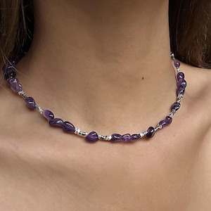May include: A necklace of polished, deep purple amethyst stones. The stones are strung with small silver-coloured beads. The necklace is worn around the neck, highlighting the natural beauty of the gemstones.