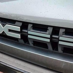 May include: The front grille of a gray vehicle featuring a black honeycomb-style mesh pattern with horizontal silver accents. Centered on the grille is a stylized silver emblem consisting of two mirrored letters forming a geometric shape.