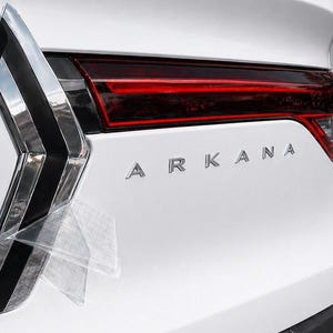 以下が含まれることがあります： 白い車両の後部で、銀色の「ARKANA」の文字が特徴です。赤い水平のテールライトアセンブリがトランク全体に広がり、左側には透明な保護フィルムが貼られた垂直のクロームと黒のライトハウジングが配置されています。