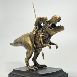May include: A gold-colored figurine of a person riding a Tyrannosaurus Rex. The person is holding a spear and is sitting on the dinosaur's back. The dinosaur is standing on a black wooden base.