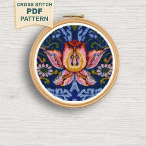 May include: A cross-stitch pattern in a wooden hoop. The design features a central pink and yellow floral motif on a dark blue background. The text "CROSS STITCH PDF PATTERN" is in a banner at the top.