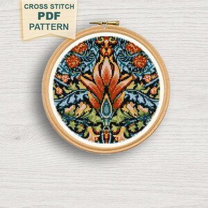 May include: A completed cross-stitch project in a wooden hoop. The design features a symmetrical floral pattern with orange, blue, green, and peach hues. The text "CROSS STITCH PDF PATTERN" is on a tan banner.