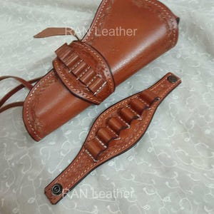 May include: A brown leather gun holster and ammunition belt. The holster features decorative stitching and a strap. The ammunition belt has loops for bullets. The text "RAN Leather" is visible.