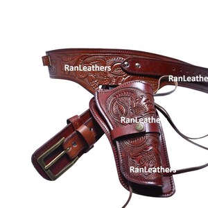 Può includere: Un set di fondina e cintura per pistola in pelle marrone con intricati motivi floreali e a foglie. Il set include una cintura con fibbia in ottone e una fondina per un'arma da fuoco. Il marchio "RanLeathers" è visibile.