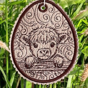 Könnte beinhalten: Ein ovaler, bestickter Anhänger mit einem Highland-Kuh-Motiv. Die Kuh ist hellbraun mit dunkelbraunen Akzenten, vor einem beigefarbenen Hintergrund mit Wirbelmustern. Der Anhänger hat einen gewellten Rand und eine Schlaufe zum Aufhängen.