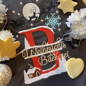 Könnte beinhalten: Ein dunkelgrauer Stoff mit einem gestickten roten Buchstaben B, einem weißen Mond, einer schwarzen Silhouette eines Schlittens mit Rentieren, einer hellblauen Schneeflocke und einer Hütte. Der Text "Weihnachts Bäckerei" ist gold gestickt. Inklusive Metall-Ausstechformen.