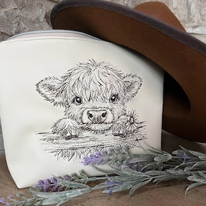 May include: Cream-coloured cosmetic bag featuring a black and white illustration of a Highland cow holding a flower. A brown cowboy hat rests behind the bag. Lavender sprigs are in the foreground.