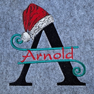 May include: Embroidered grey fabric featuring a black letter "A" adorned with a red Santa hat and white trim. The name "Arnold" is embroidered in red, with teal flourishes.